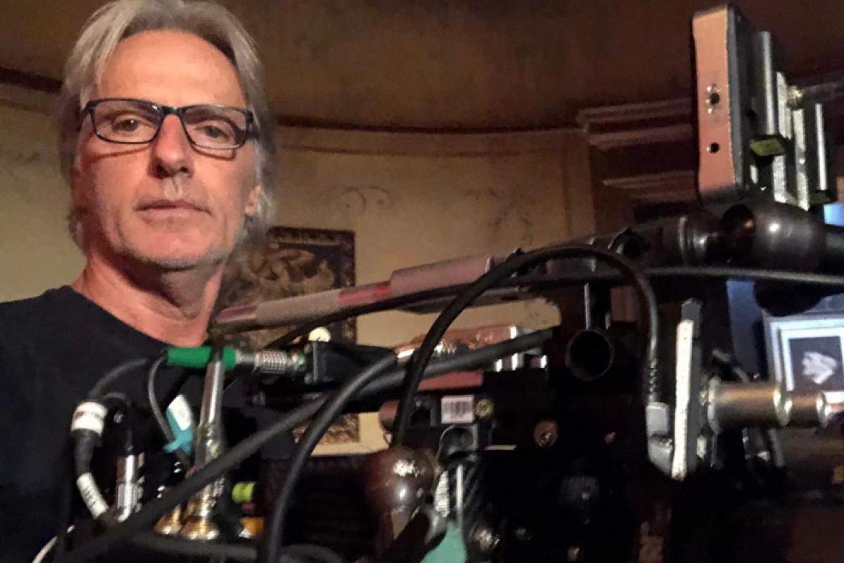 Homem de cabelos grisalhos e óculos, vestido com camiseta preta, ao lado de uma câmera cinematográfica em um ambiente interno de paredes decoradas.